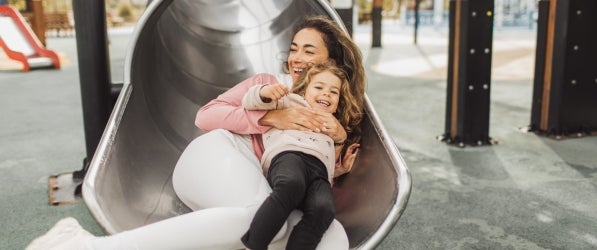 A woman holding a child going down a playground slide, both smiling.